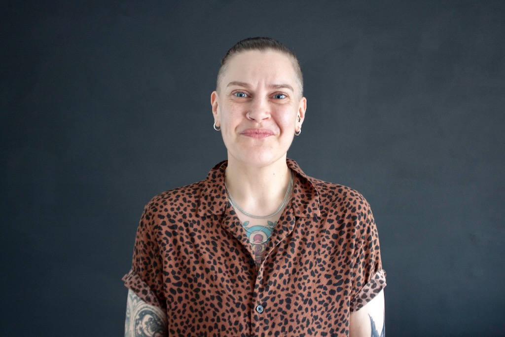 Person with tattoos smiling in front of grey wall